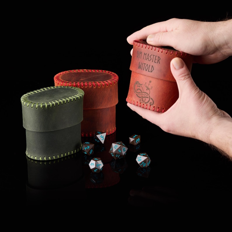 May include: Two cylindrical leather dice cases in olive green and reddish-brown with visible stitching along the edges. The reddish-brown case features the text DM MASTER WITOLD and a dragon emblem. Several multi-sided dice with blue numbers are scattered.