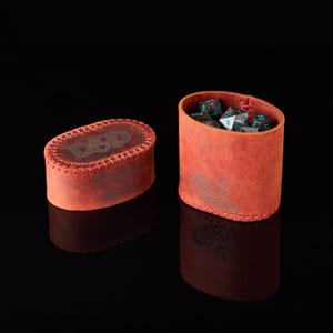 May include: An oval-shaped red leather dice container with a matching lid, featuring visible red stitching along the edges. The lid displays the D&D logo, while the side of the container shows a dragon emblem. Inside are several multi-sided dice.