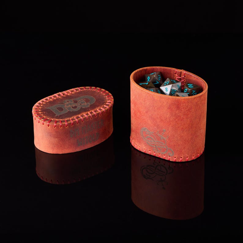 May include: An oval-shaped red leather dice container with a matching lid, featuring visible red stitching along the edges. The lid displays the D&D logo, while the side of the container shows a dragon emblem. Inside are several multi-sided dice.