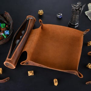 May include: A brown leather dice tray with a matching leather dice rolling tube. The tray has a flap that folds over to create a closed container. The tube has a zipper closure. There are several dice scattered around the tray and tube.
