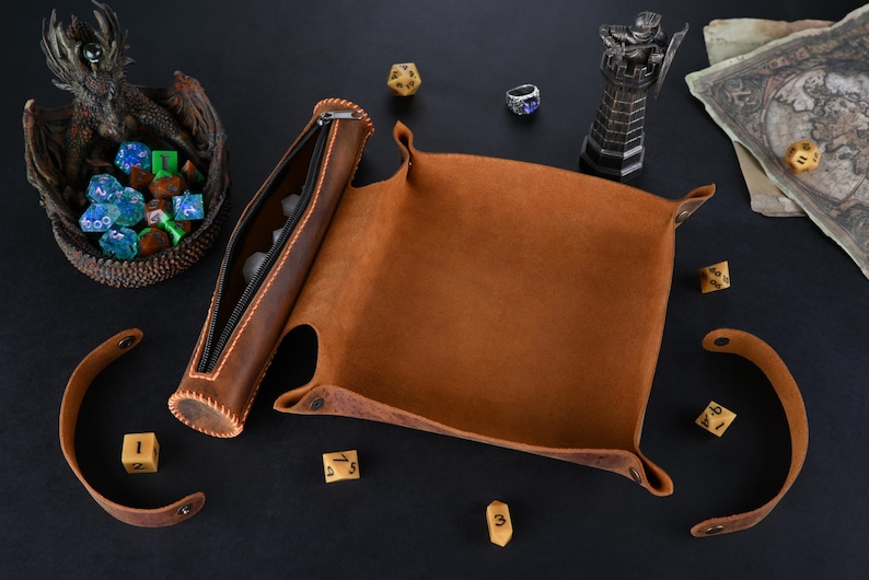 May include: A brown leather dice tray with a matching leather dice rolling tube. The tray has a flap that folds over to create a closed container. The tube has a zipper closure. There are several dice scattered around the tray and tube.