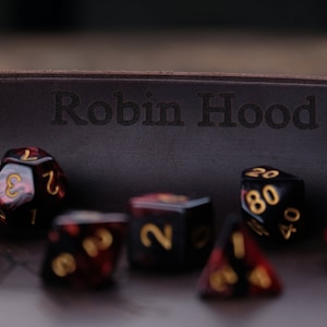 May include: A brown leather dice tray with the words "Robin Hood" engraved on the front. The tray is filled with red and black dice with gold numbers.