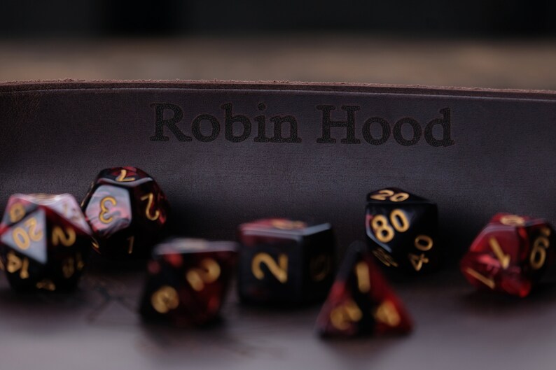 May include: A brown leather dice tray with the words "Robin Hood" engraved on the front. The tray is filled with red and black dice with gold numbers.