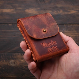 Puede incluir: Un estuche para tarjetas de cuero marr&oacute;n con el texto "Uncle Ace" grabado en la parte delantera.