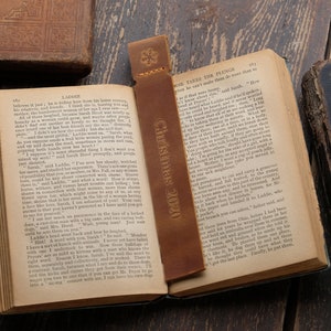 Pode incluir: Um marcador de p&aacute;gina de couro castanho com o texto "Christmas 2020" nele. O marcador est&aacute; colocado em um livro aberto com texto nas p&aacute;ginas. O livro est&aacute; sobre uma superf&iacute;cie de madeira.