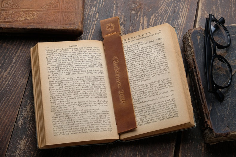 Pode incluir: Um marcador de p&aacute;gina de couro castanho com o texto "Christmas 2020" nele. O marcador est&aacute; colocado em um livro aberto com texto nas p&aacute;ginas. O livro est&aacute; sobre uma superf&iacute;cie de madeira.
