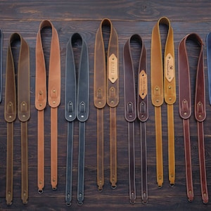 Puede incluir: Una colección de 12 correas de cámara de cuero en varios colores, incluyendo marrón, beige, negro y azul. Cada correa tiene un borde cosido y una pequeña etiqueta de cuero con un nombre en ella.