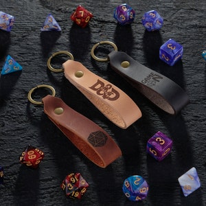 May include: Three leather keychains with different designs. The keychains are brown, black, and tan. The brown keychain has a D&D logo, the black keychain has the text "GAME ON", and the tan keychain has a dice symbol. The keychains are surrounded by various colored dice.