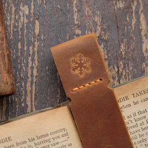 Pode incluir: Um marcador de p&aacute;gina de couro marrom com um design de floco de neve. O marcador est&aacute; dobrado ao meio e est&aacute; em cima de um livro aberto. O texto do livro diz "Laddie Takes the Plunge".