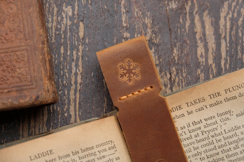 Pode incluir: Um marcador de p&aacute;gina de couro marrom com um design de floco de neve. O marcador est&aacute; dobrado ao meio e est&aacute; em cima de um livro aberto. O texto do livro diz "Laddie Takes the Plunge".