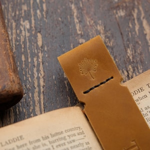 Pode incluir: Um marcador de p&aacute;gina de couro castanho com uma borda costurada e um pequeno desenho de flor em relevo. O marcador est&aacute; sobre um livro aberto com o texto "LADDIE" e "ODIE TAKES THE PI" vis&iacute;vel.