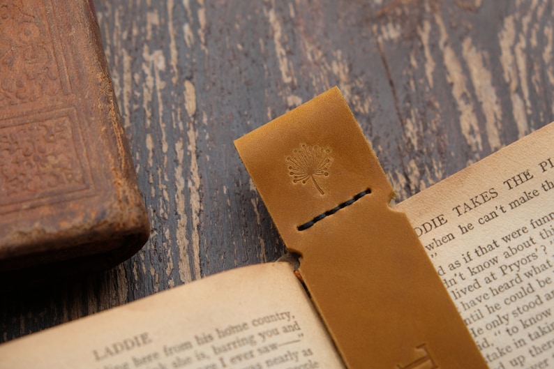 Pode incluir: Um marcador de p&aacute;gina de couro castanho com uma borda costurada e um pequeno desenho de flor em relevo. O marcador est&aacute; sobre um livro aberto com o texto "LADDIE" e "ODIE TAKES THE PI" vis&iacute;vel.