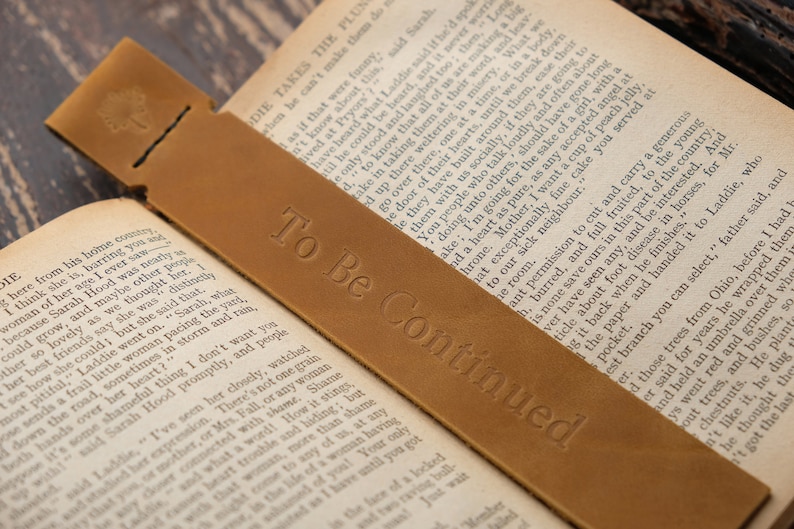 Pode incluir: Um marcador de p&aacute;gina de couro castanho com o texto "To Be Continued" gravado em relevo. O marcador est&aacute; sobre um livro aberto.