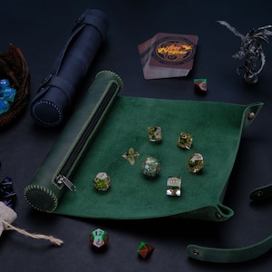 May include: A green leather dice tray with a matching leather dice rolling tube. The tray has a green felt lining and is decorated with stitching. The tube has a zipper closure. There are several dice on the tray and tube, including a set of green and gold dice.