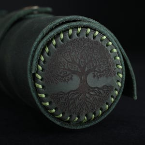 May include: A dark green leather roll with a light green stitched border. The roll has a tree of life design embossed in the center.