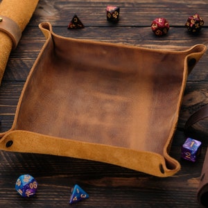 May include: A brown leather dice tray with a suede interior. The tray is surrounded by various colored dice on a wooden surface.