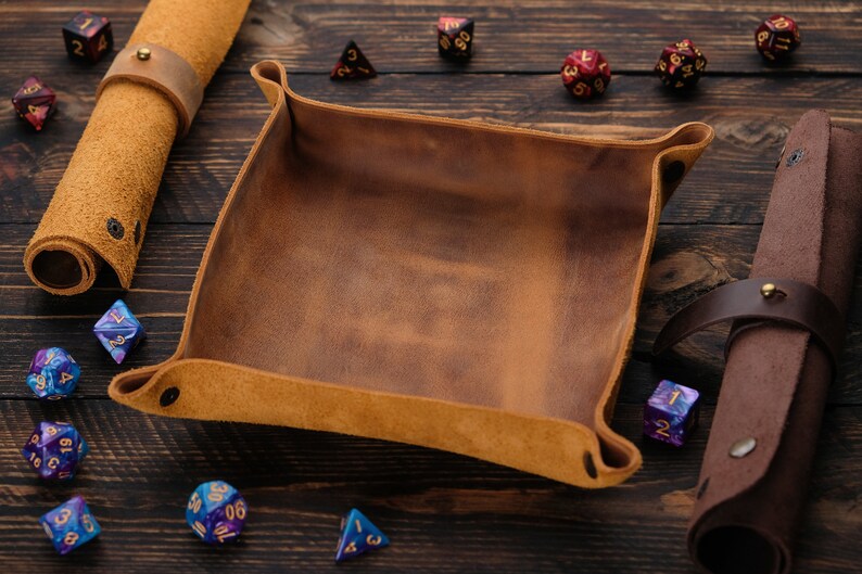 May include: A brown leather dice tray with a suede interior. The tray is surrounded by various colored dice on a wooden surface.