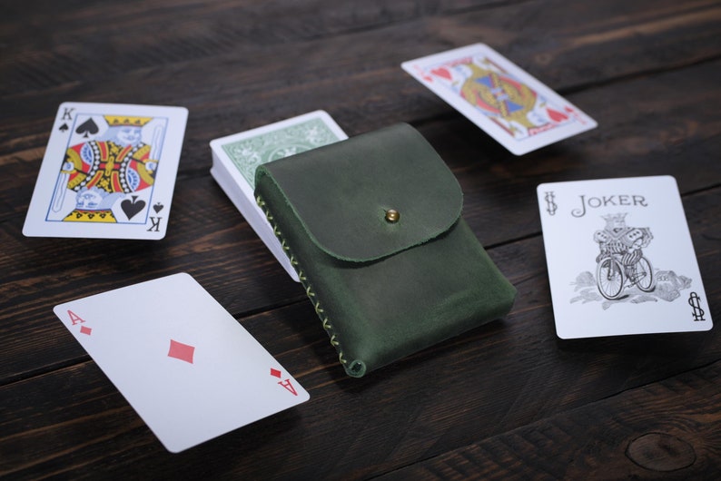 Puede incluir: Un estuche para cartas de cuero verde con una solapa y un cierre de bot&oacute;n de lat&oacute;n. El estuche contiene una baraja de cartas. Hay cuatro cartas en la mesa: el rey de picas, el as de diamantes, la reina de corazones y el comod&iacute;n.