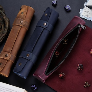 Könnte beinhalten: Drei Würfelbehälter aus Leder in Braun, Marineblau und Burgunderrot. Die braunen und marineblauen Etuis sind aufgerollt und mit Riemen befestigt. Das burgunderrote Etui ist geöffnet und zeigt Würfel im Inneren. Eine Drachenfigur und Würfel sind im Hintergrund.