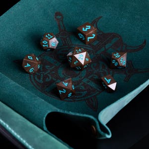 Pu&ograve; includere: Un set di dadi poliedrici marroni con numeri turchese, appoggiati su una superficie di feltro verde acqua. I dadi sono disposti su un tappetino da gioco con un design grafico nero. Per giochi di ruolo.