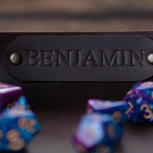 May include: A brown leather dice tray with the name "BENJAMIN" embossed on the front. The tray is filled with colorful dice.