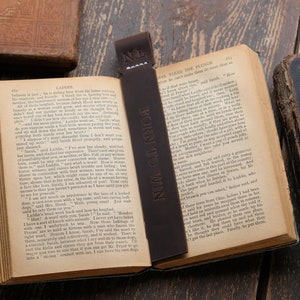 Pode incluir: Um marcador de p&aacute;gina de couro castanho com o texto "BORN TO WIN" em relevo. O marcador est&aacute; colocado num livro aberto com as p&aacute;ginas viradas para um cap&iacute;tulo intitulado "LADGRE".