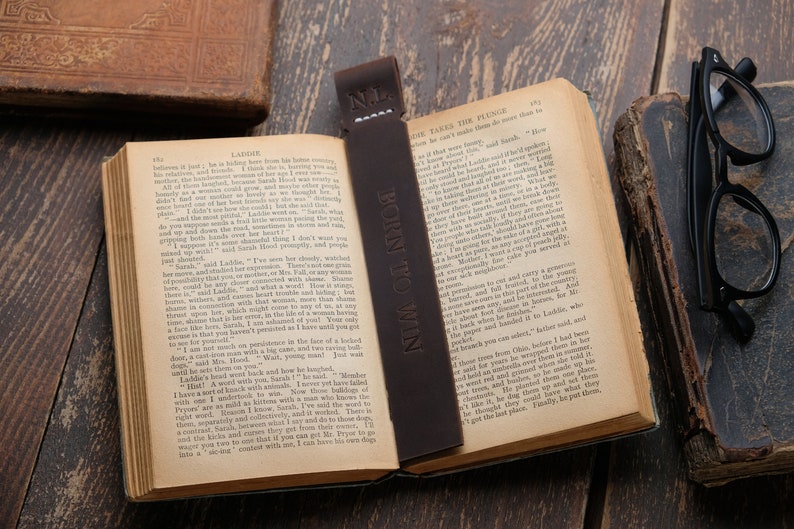Pode incluir: Um marcador de p&aacute;gina de couro castanho com o texto "BORN TO WIN" em relevo. O marcador est&aacute; colocado num livro aberto com as p&aacute;ginas viradas para um cap&iacute;tulo intitulado "LADGRE".