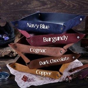 May include: A stack of five leather dice trays in different colors: navy blue, burgundy, cognac, dark chocolate, and copper. Each tray has a different color and is labeled with its color name.