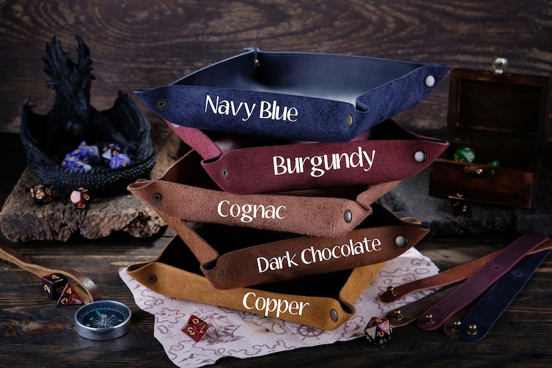 May include: A stack of five leather dice trays in different colors: navy blue, burgundy, cognac, dark chocolate, and copper. Each tray has a different color and is labeled with its color name.