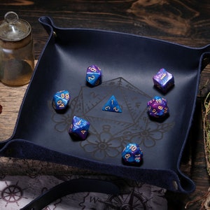 May include: A blue felt dice tray with a black and white design featuring a pentagram and floral motifs. The tray contains six iridescent dice with gold numbers.