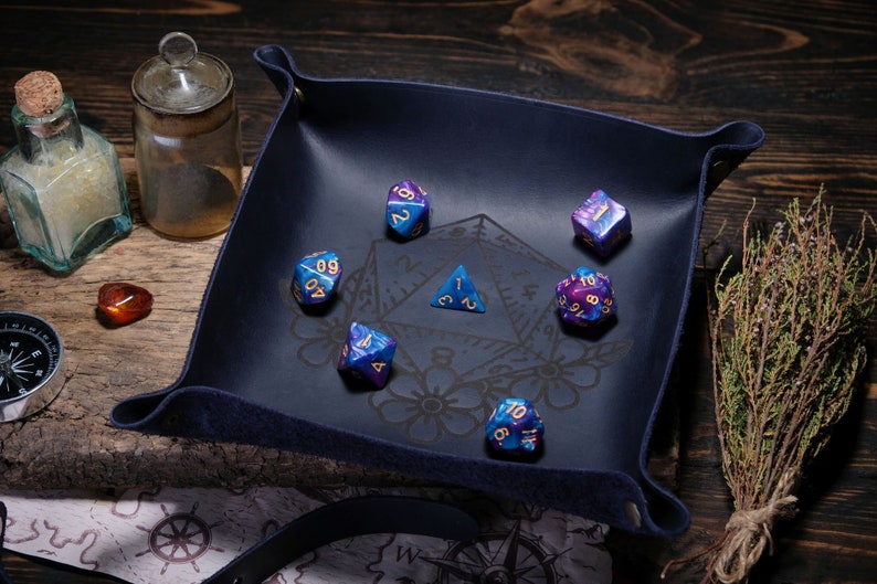 May include: A blue felt dice tray with a black and white design featuring a pentagram and floral motifs. The tray contains six iridescent dice with gold numbers.