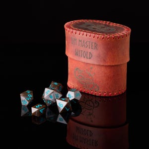 May include: A cylindrical brown leather dice case with red stitching, featuring the embossed text DM MASTER WITOLD and a dragon graphic. Beside the case are several polyhedral dice with dark brown surfaces and bright cyan numbers for gaming.
