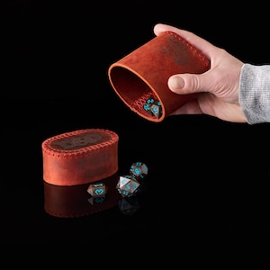 May include: A cylindrical red leather dice cup with red stitching and a matching oval lid, both embossed with D&D branding. Several dark brown polyhedral dice with bright blue numbers are scattered on the surface and inside the cup.