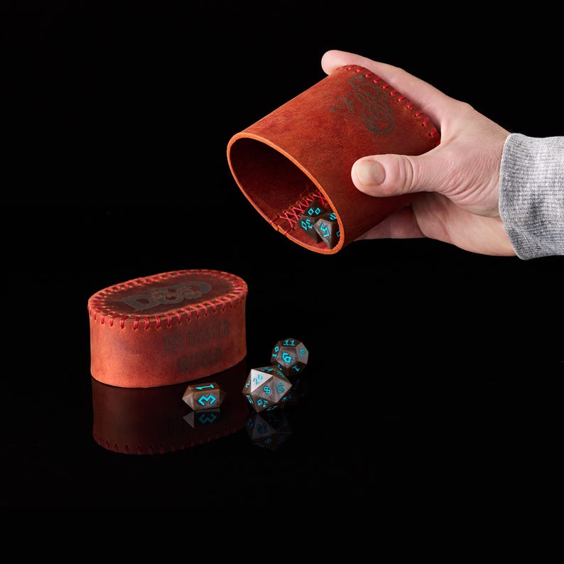 May include: A cylindrical red leather dice cup with red stitching and a matching oval lid, both embossed with D&D branding. Several dark brown polyhedral dice with bright blue numbers are scattered on the surface and inside the cup.