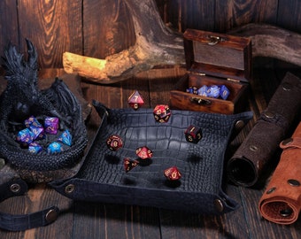 May include: A black leather dice tray with a crocodile pattern, with red and blue dice inside and scattered around it. There is a black dragon statue with blue and red dice inside, a wooden box with blue dice inside, and two leather dice rolling pouches.