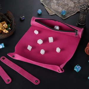 Könnte beinhalten: Ein rosa Leder-Würfeltablett mit passendem Würfelturm und vier Würfelhaltern. Das Tablett hat einen Reißverschluss und lässt sich zum Verstauen zusammenrollen.