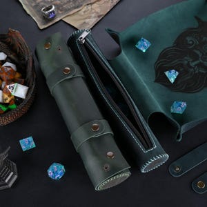 May include: A green leather dice rolling tray with a black and white illustration of a creature's face. The tray is rolled up and secured with a zipper. There is a matching green leather dice bag with a zipper and a matching green leather strap.