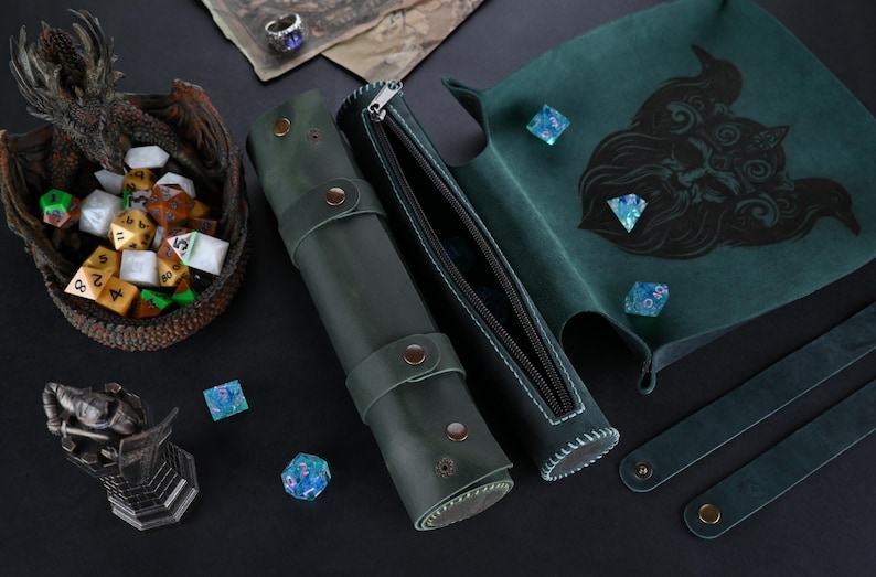 May include: A green leather dice rolling tray with a black and white illustration of a creature's face. The tray is rolled up and secured with a zipper. There is a matching green leather dice bag with a zipper and a matching green leather strap.