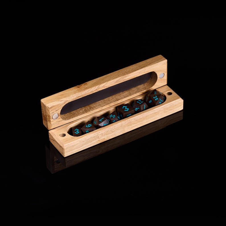 Pu&ograve; includere: Una scatola di dadi in legno chiaro, aperta per rivelare un set di dadi marrone scuro con segni turchesi. La scatola ha una chiusura magnetica e una forma rettangolare. I dadi sono ordinatamente disposti all'interno della scatola.