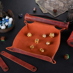 May include: A brown leather dice tray with a red leather roll-up dice bag. The tray has a red felt lining and is filled with gold dice. The dice bag has a sun design on the front.