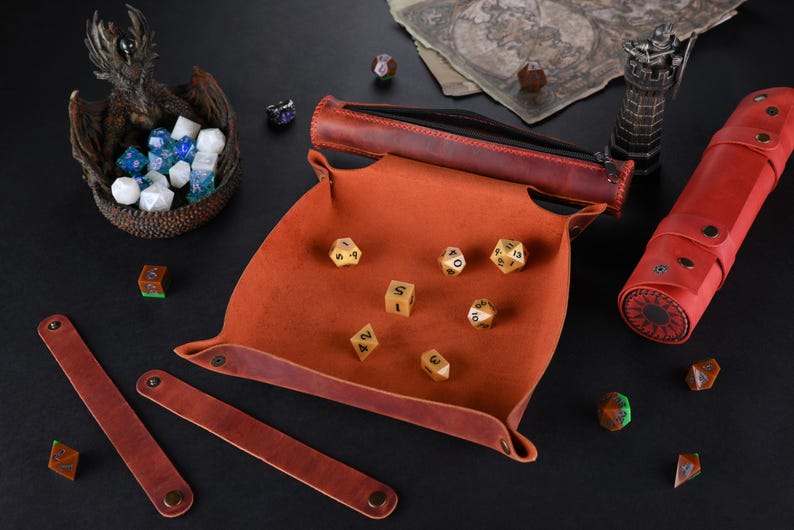 May include: A brown leather dice tray with a red leather roll-up dice bag. The tray has a red felt lining and is filled with gold dice. The dice bag has a sun design on the front.