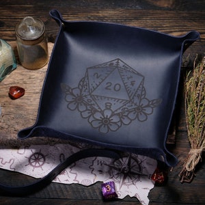 May include: A dark blue leather dice tray with a floral design and a 20-sided die engraved in the center. The tray is surrounded by a compass, a map, and other gaming accessories.