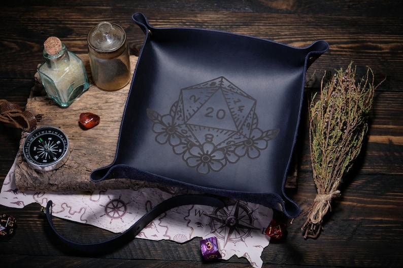 May include: A dark blue leather dice tray with a floral design and a 20-sided die engraved in the center. The tray is surrounded by a compass, a map, and other gaming accessories.