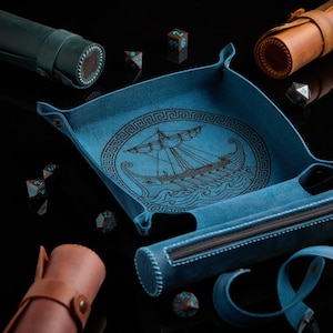 May include: A blue leather dice tray featuring a black embossed ancient Greek ship design surrounded by a geometric border. The tray includes an integrated zippered storage compartment and is accompanied by several multi-sided dice and leather cases.