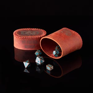 May include: A reddish-brown leather dice cup with a matching lid, featuring stitched edges and the embossed text D&D and DM MASTER WORLD. Several dark-colored polyhedral dice with bright blue numbers are scattered in front of the open cup.