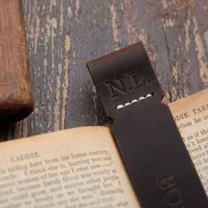 Pode incluir: Um marcador de p&aacute;gina de couro castanho com as iniciais "N.L." gravadas em branco. O marcador de p&aacute;gina est&aacute; colocado num livro com o texto "Laddie Takes The Plunge" vis&iacute;vel.