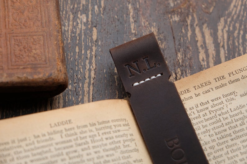 Pode incluir: Um marcador de p&aacute;gina de couro castanho com as iniciais "N.L." gravadas em branco. O marcador de p&aacute;gina est&aacute; colocado num livro com o texto "Laddie Takes The Plunge" vis&iacute;vel.