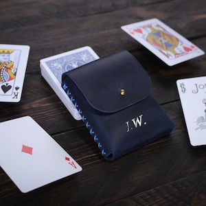Puede incluir: Un estuche para tarjetas de cuero azul con detalles de costura azul y las iniciales "J.W." grabadas en la parte delantera. El estuche est&aacute; abierto y se puede ver una baraja de cartas en el interior. Tambi&eacute;n hay varias cartas de juego esparcidas sobre una superficie de madera.