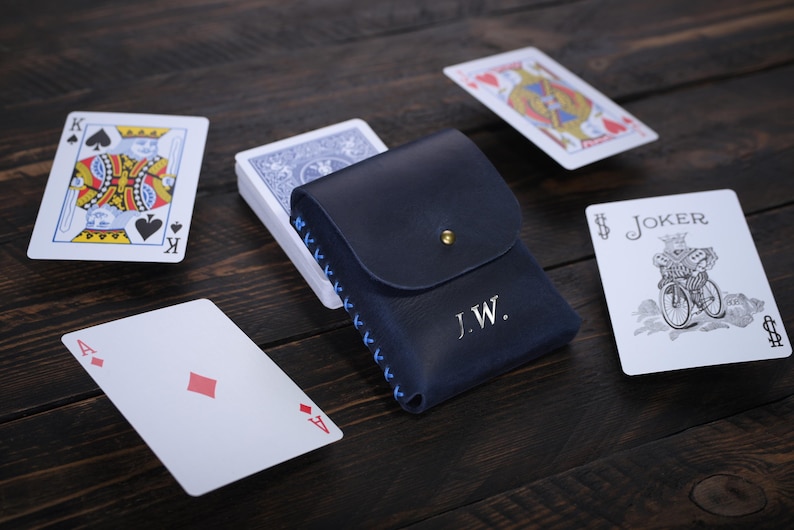Puede incluir: Un estuche para tarjetas de cuero azul con detalles de costura azul y las iniciales "J.W." grabadas en la parte delantera. El estuche est&aacute; abierto y se puede ver una baraja de cartas en el interior. Tambi&eacute;n hay varias cartas de juego esparcidas sobre una superficie de madera.