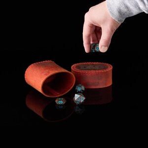 May include: A set of dark brown polyhedral dice with bright blue numbers, accompanied by a cylindrical orange leather dice cup and a matching oval leather case featuring the embossed text D&D and DM MASTER SHIELD on the lid.